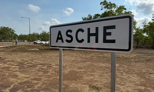 Roadside sign reading 'Asche' in a dry, grassy area with trees and parked cars in the background.